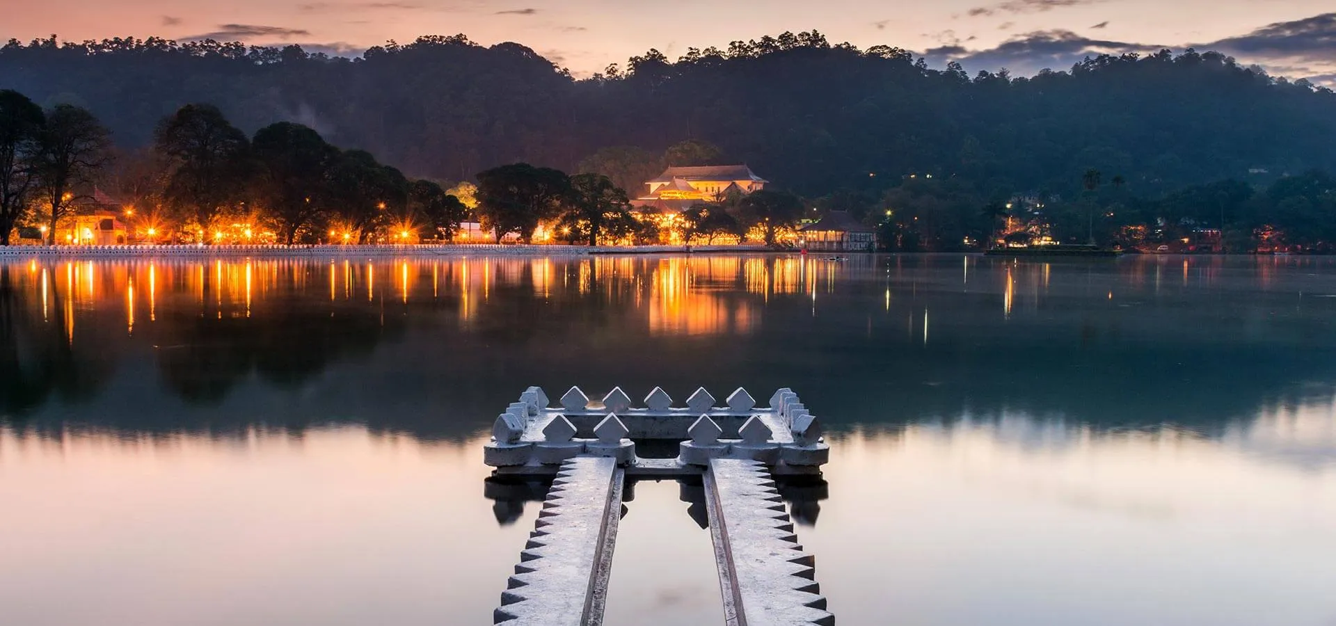 Kandy Lake at dusk with shoreline lights reflected in the water and forested hills beyond